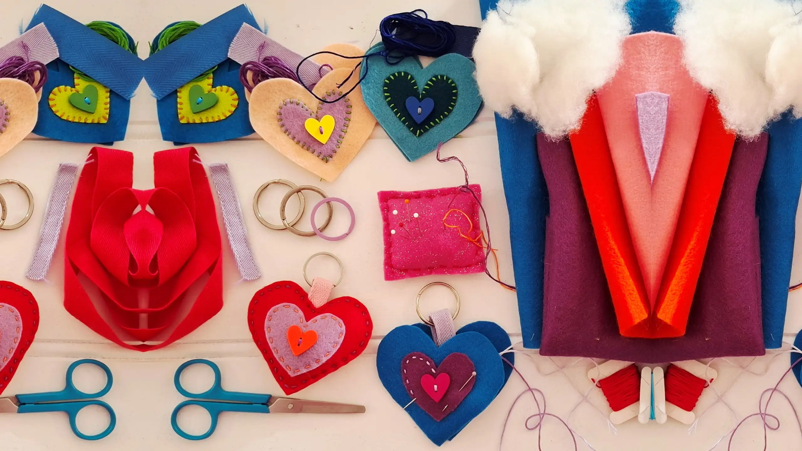 Colorful handmade felt crafts displayed on a table, including heart-shaped keyrings, stitched hanging decorations, a pincushion, scissors, thread, and bright folded felt projects.